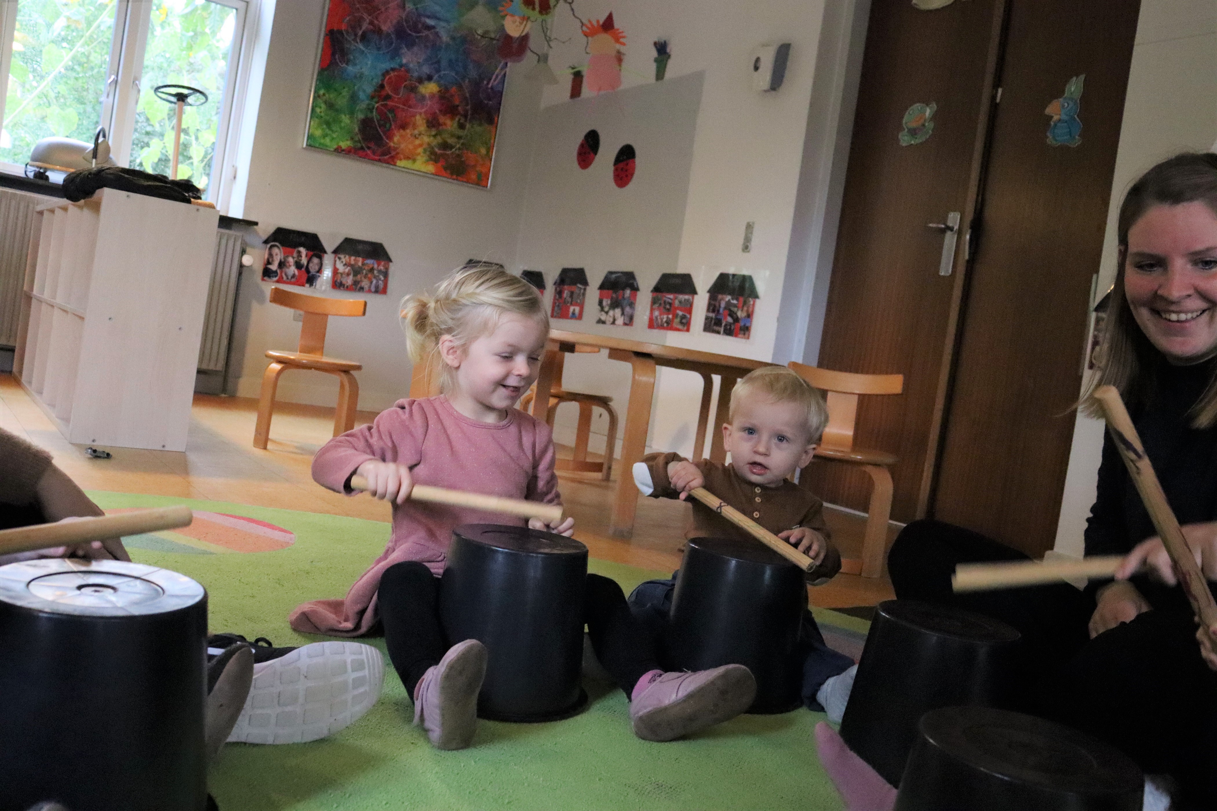 Samling hvor pædagog og børn spiller på små spande med trommestikker
